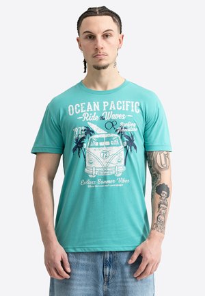 Mladý muž s vrkočmi, ktorý má na sebe tyrkysové tričko Ocean Pacific s potlačou a svetlomodré džínsy, ukazuje tetovania na pravej ruke.
