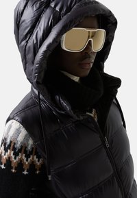 Schwarze Steppweste mit glänzender Textur und gepolstertem Design; verfügt über eine große Kapuze. Zubehör umfasst übergroße hellfarbige Sonnenbrillen.