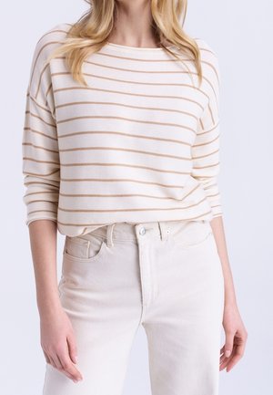 Femme portant un haut blanc à manches longues avec de fines rayures horizontales beiges, rentré dans un jean taille haute écru.