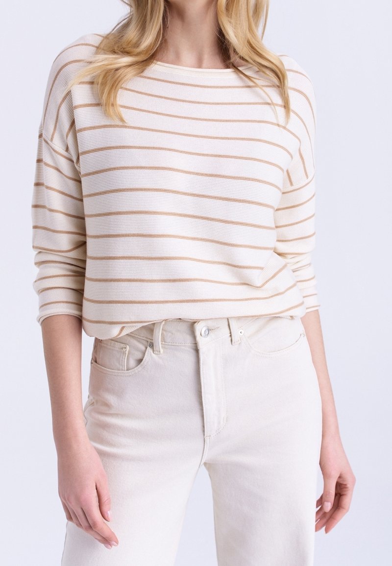 Femme portant un haut blanc à manches longues avec de fines rayures horizontales beiges, rentré dans un jean taille haute écru.