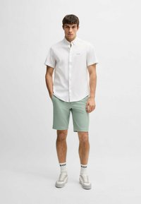 White button-up shirt with short sleeves, paired with light green shorts. Model wears grey sneakers and white socks with logo accents.