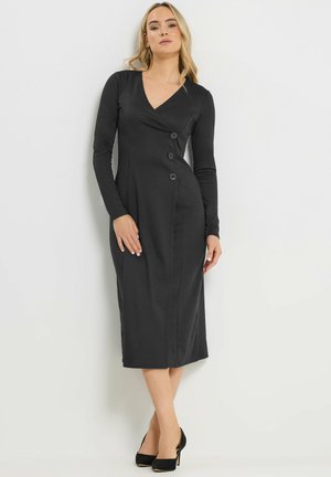 Vrouw draagt een zwarte midi-jurk met lange mouwen en drie knopen aan de voorkant, gecombineerd met zwarte pumps, staand tegen een witte muur.