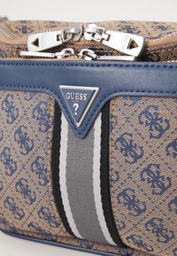 Bolso de tela marrón con estampado y un acento de cuero azul, que cuenta con un cierre de plata y un prominente logo triangular. Dos franjas en gris y blanco.