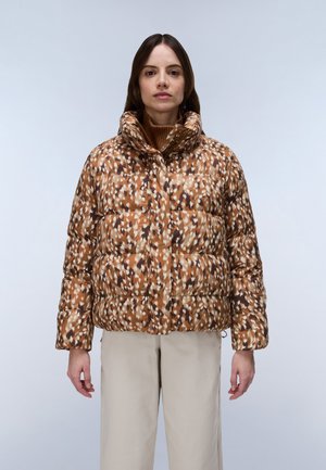 Femme portant une doudoune marron à motifs avec un col montant et un pantalon beige, debout devant un fond clair uni.