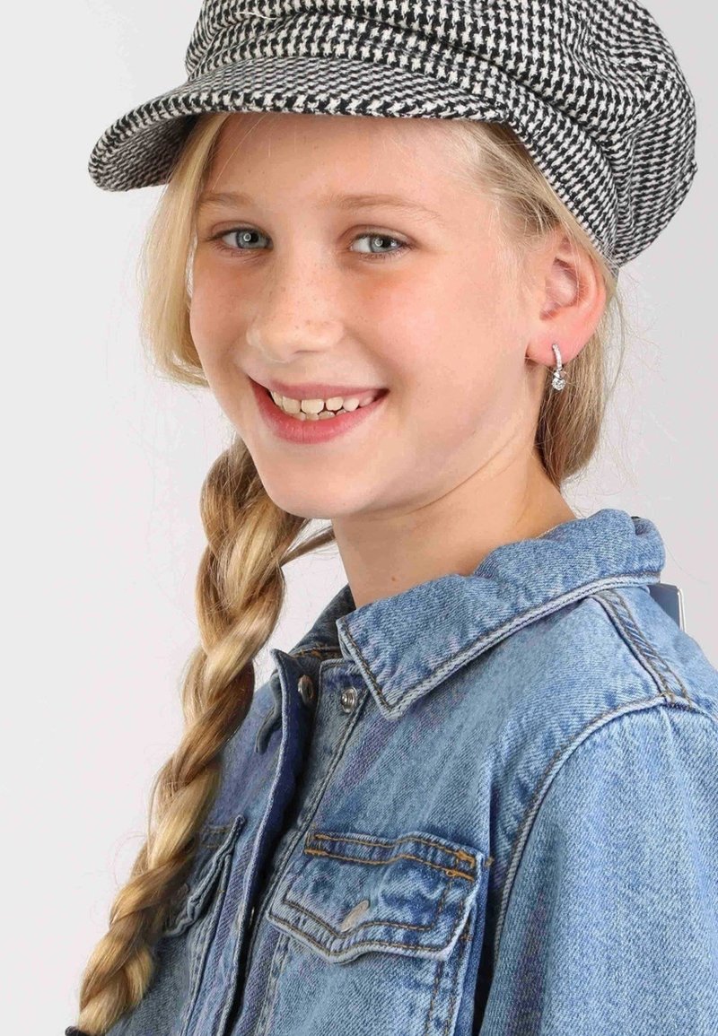 Casquette en noir et blanc à motif pied-de-poule, veste en denim avec fermeture à boutons, tresse blond clair et petites boucles d'oreilles scintillantes.