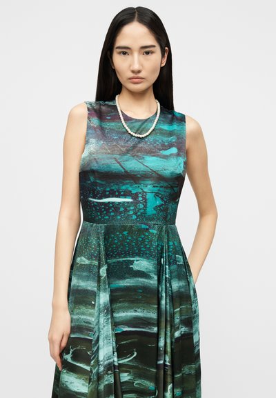 Γυναίκα με αμάνικο φόρεμα σε τιρκουάζ και μαύρο με σχέδια, που φορά μαργαριταρένιο κολιέ, στέκεται μπροστά σε απλό φωτεινό φόντο.