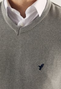 Szary dzianinowy sweter z dekoltem w serek, z prążkowanym wykończeniem i małym granatowym logo ptaka na piersi. Nałożony na białą koszulę.