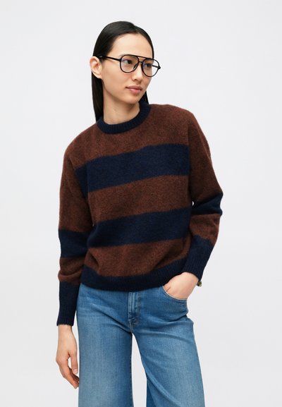 Pulover tricotat cu dungi maro și bleumarin, cu guler rotund, confecționat dintr-un material moale. Asortat cu blugi albastre deschis, cu croială lejeră.