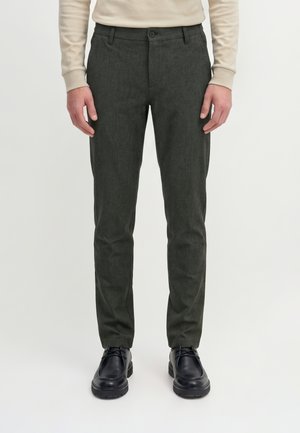 Mann trägt dunkelgraue Slim-Fit-Hose, beiges langärmeliges Hemd und schwarze Schnürschuhe, steht vor einfachem hellem Hintergrund.