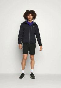 Μαύρο φερμουάρ μπουφάν με ψηλό κολάρο, γκρίζα εσωτερική επένδυση και λογότυπο CK. Συνδυασμένο με μαύρα σορτς και αθλητικά παπούτσια, σε στάση μπροστά από ουδέτερη φόντο.