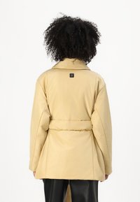 Beige pufjakke med oversized pasform, bred krave og bæltesyning i taljen. Har et logo-patch bagpå og en glat tekstur.