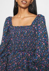 Kukkamekko, jossa on neliön muotoinen pääntie, kerätty smokkitekniikka, pitkät puffihihat ja tummansininen tausta, jossa on vaaleanpunaisia ja sinisiä kukkakuviota.