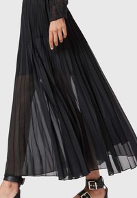 Gonna nera plissettata in tessuto trasparente, caratterizzata da pieghe verticali, un design fluido e un taglio alla caviglia. Accompagnata da un sandalo nero con cinturini.