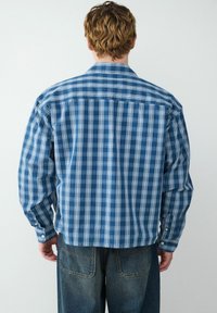 Camicia a quadri blu con maniche lunghe, caratterizzata da strisce bianche. Il materiale sembra leggero, con una vestibilità casual e orlo arrotondato.