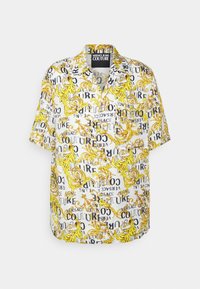 Valkoinen lyhythihainen paita, jossa on keltaisia barokkikierteitä ja musta teksti, joka lukee "Versace Jeans Couture" eri asennoissa, etutasku näkyvissä.