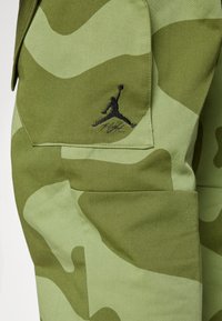 Kamufliažo krovinių kelnės iš šviesiai ir tamsiai žalios medžiagos, su juodu siuvinėtu Jumpman logotipu ir "Flight" tekstu ant kišenės.