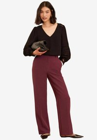 Fekete blúz V-kivágással és csipke díszítéssel, magas derékú bordó, bőszárú nadrággal párosítva. A modellen fekete clutch található.