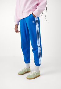 Pantaloni della tuta blu con strisce bianche laterali e logo adidas. Abbinati a scarpe da ginnastica verdi, calzini bianchi e una felpa rosa chiaro.