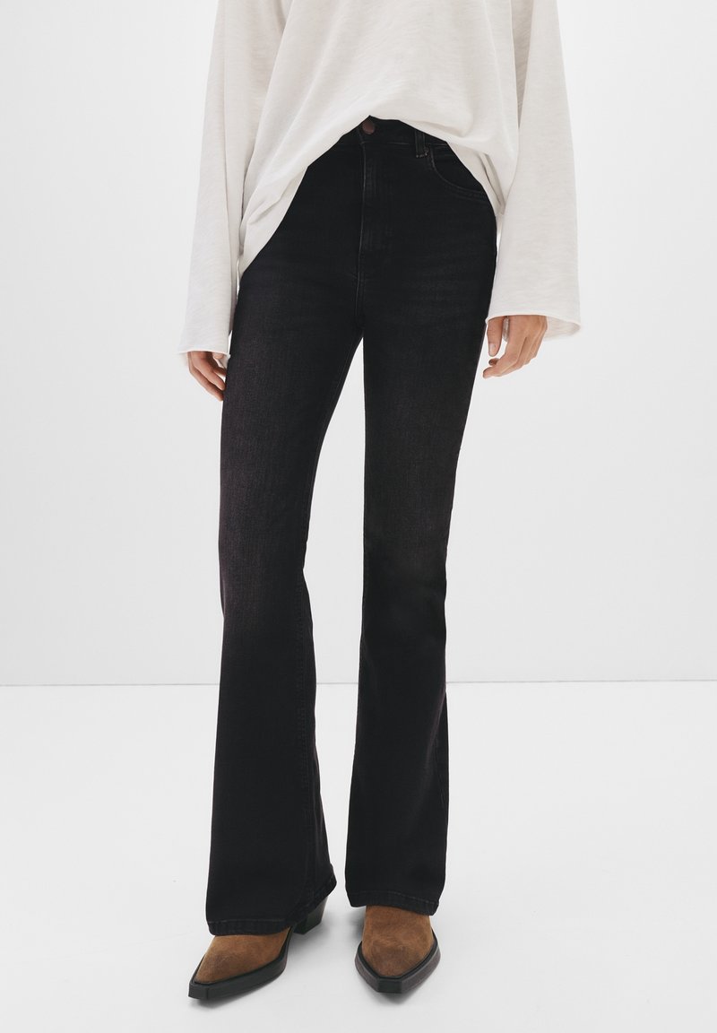 Jean évasé noir en denim à taille haute, présentant une texture lisse et des détails de couture minimaux, associé à des bottes marron à bout pointu.