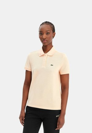 Kvinde iført en lyse ferskenfarvet kortærmet polo med en lille grøn logo og sorte bukser, stående mod en ensfarvet baggrund.