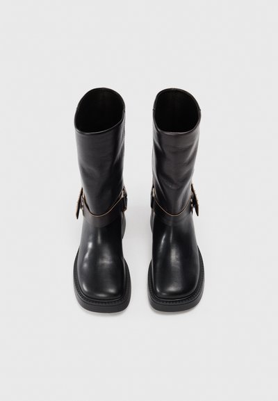 Bottines en cuir noir avec un bout carré, texture lisse, une sangle marron contrastante avec un détail de boucle en or, et une semelle en caoutchouc plate.