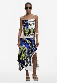 Multifarvet trykt kjole med en tætsiddende top, asymmetrisk kant, dristige blå og gule grafikker samt hvide accenter; kombineret med platformsandaler.