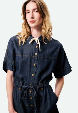 Femme aux cheveux blonds ondulés portant une combinaison en jean foncé à manches courtes, avec des boutons sur le devant et une taille nouée, debout devant un fond blanc.