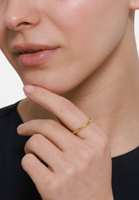 Złota skręcona obrączka noszona na palcu, o gładkiej i odbijającej światło powierzchni w zestawieniu z neutralnym odcieniem skóry, doskonale uzupełniająca minimalistyczny czarny top.
