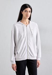Sweatshirt cinza claro com fecho e tecido macio, apresentando capuz com cordão, acessórios dourados e bolsos laterais, combinado com calças pretas.
