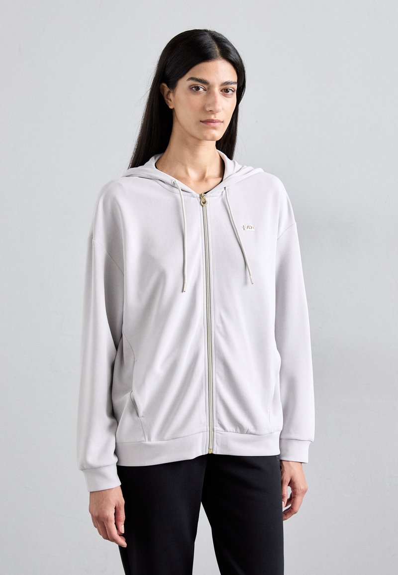 Sweatshirt cinza claro com fecho e tecido macio, apresentando capuz com cordão, acessórios dourados e bolsos laterais, combinado com calças pretas.