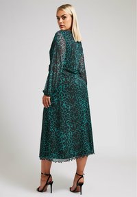 Transparente tealfarbene Kleid mit schwarzem Leopardenmuster, ausgestattet mit langen Ärmeln und einem wellenförmigen Saum. Kombiniert mit schwarzen, geschnürten High Heels.