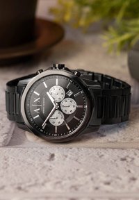 Musta kronograaf kell tekstureeritud musta silikoonrihmaga, hõbedaste aktsentidega, kolme aladialiga ja silmapaistva "AX" logoga kellanäidikul.