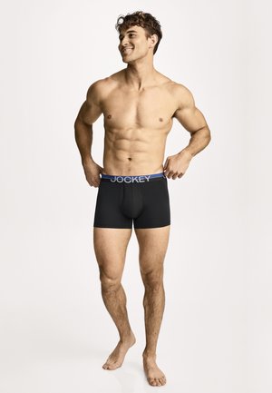 Lächelnder muskulöser Mann, der ein schwarzes Jockey-Boxershorts mit blauem Bund anpasst und barfuß vor einem schlichten hellen Hintergrund steht.