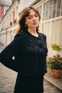 Noor naine laineliste juustega kannab musta sädelevat kampsunit ja seelikut, seisab munakivisel tänaval hoonete ja lillepottide lähedal.