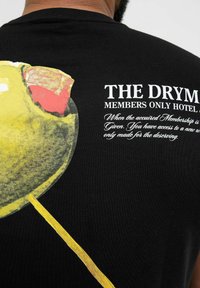 Schwarzes T-Shirt mit einem bunten Grafikdesign eines Lutschers, mit gelbem Stiel und roten, grünen sowie pinken Akzenten. Der Text lautet „THE DRY MI...“ in Weiß.