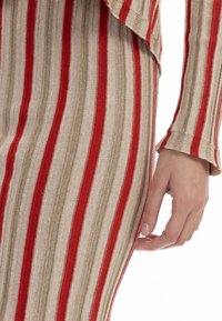Rochie rayee cu un material texturat roșu, bej și auriu, cu un design nervurat și un mânec flared. Prim-plan al unei mâini pe articolul vestimentar.