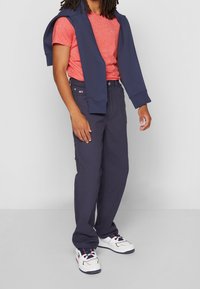 Persona vestida con una camiseta color coral, pantalones azul marino, zapatillas blancas, con un sudadera azul marino sobre los hombros.