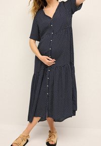 Laivansiniset napitettava äitiysmekko, jossa on valkoisia pilkkuja, lyhyet hihat ja kerroksellinen helma. Yhdistetty vaaleanruskeisiin sandaaleihin.