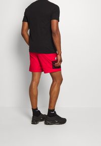 Chemise noire à manches courtes, short rouge avec poche en mesh sur le côté et chaussures de sport noires avec amortissement à air ; design minimal, look sportif.
