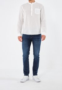 Camicia bianca di lino a maniche lunghe con colletto alla cinese e una tasca sul petto, abbinata a jeans blu scuro e sneakers bianche.