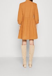 Abito in velluto a coste marrone con design svasato, maniche puff a tre quarti e vita cinchata, abbinato a stivali al ginocchio in suede beige.