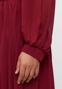 Burgundy dress with long, puffed sleeves and elastic cuffs. Smooth, flowing fabric drapes softly, with waistband gathering for shape and fit.