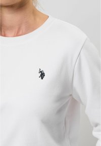 Balta džemperis ar apaļu kakla izgriezumu, kurā ir mazs melns izšūts logo uz krūtīm. Izgatavots no mīkstas, teksturētas auduma.