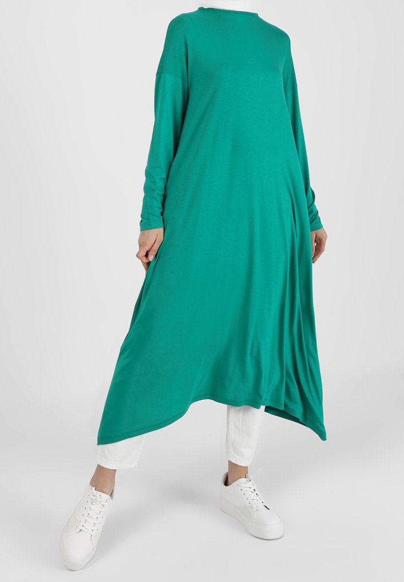 Robe longue verte en tissu doux avec une coupe décontractée, encolure arrondie et épaules tombantes, associée à des baskets blanches.
