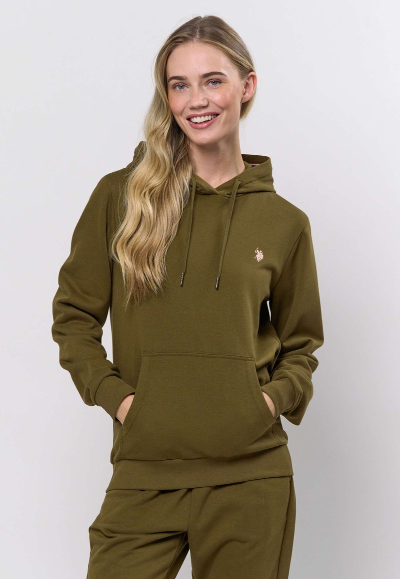 Femme souriante aux longs cheveux blonds portant un sweat à capuche vert olive avec des poches avant et un pantalon assorti sur un fond uni.