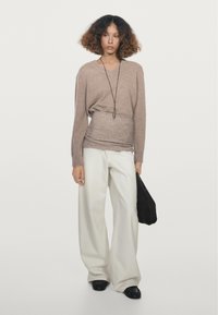 Kvinde med krøllet hår iført en beige sweater, hvide brede bukser, sorte sko og holder en sort taske mod en ensartet baggrund.