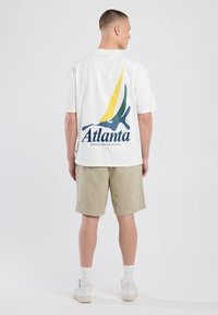 Camiseta blanca de algodón con un gráfico de un velero colorido y el texto "Atlanta" en la parte posterior. Combinada con pantalones cortos beige y zapatillas blancas.