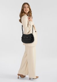 Borsa a tracolla in pelle nera con doppia patta, finiture argentate e tracolla regolabile, indossata da una modella con un completo chiaro e tacchi.