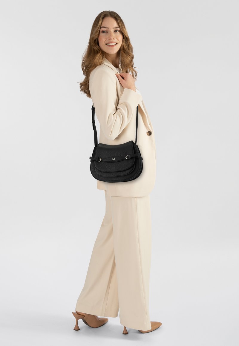 Borsa a tracolla in pelle nera con doppia patta, finiture argentate e tracolla regolabile, indossata da una modella con un completo chiaro e tacchi.