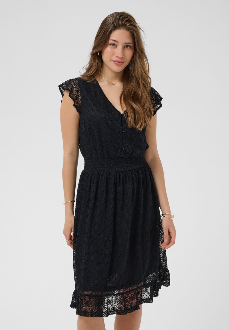 Jeune femme aux longs cheveux bruns portant une robe noire en dentelle longueur genou avec des manches courtes, debout devant un fond clair uni.